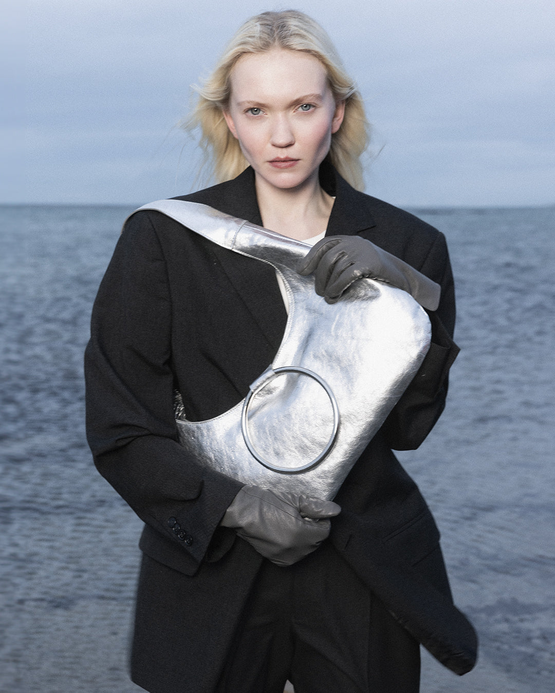A person holding a KALDA silver bag with a signature metal circle closure, standing by a body of water.