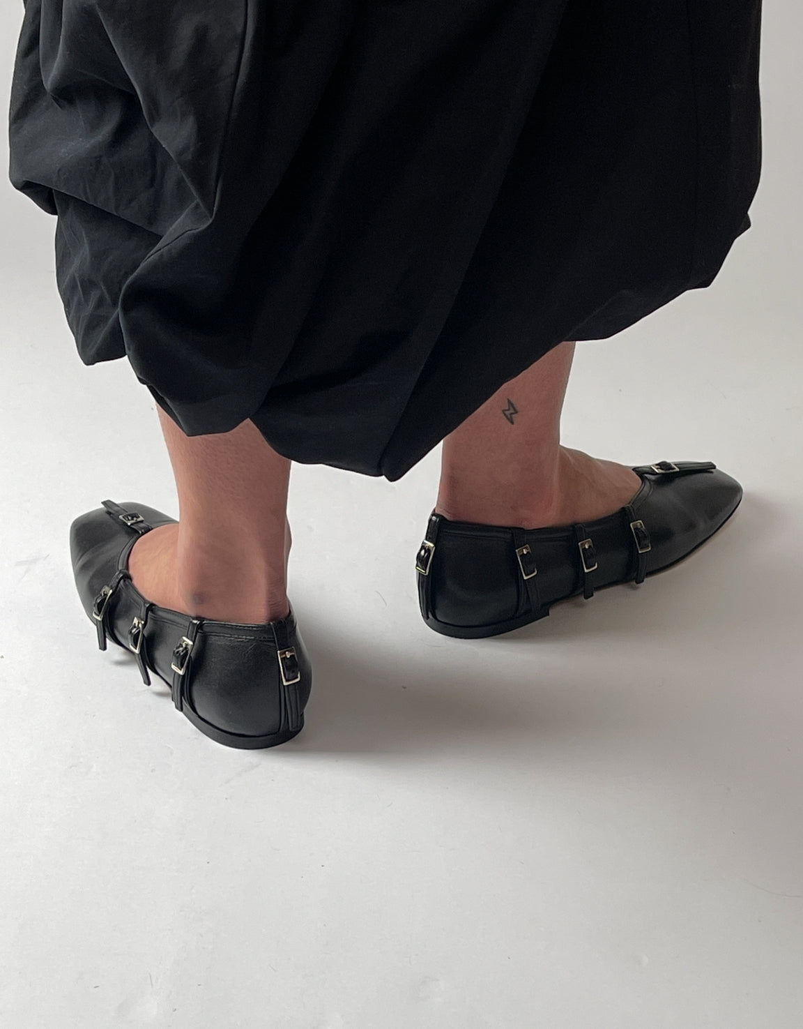 Black flat shoes with buckles worn by a person on a white background