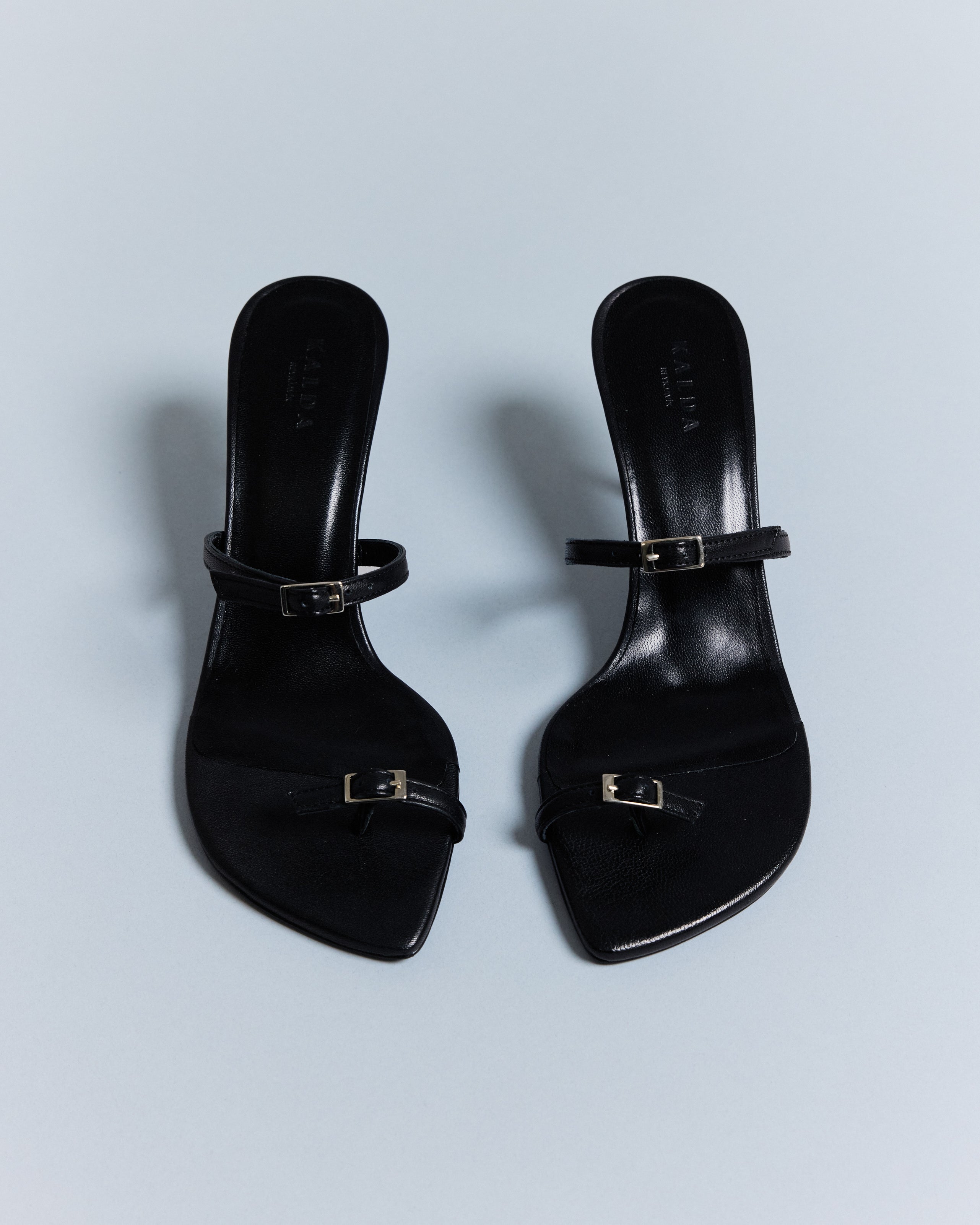 A pair of KALDA black summer sandals with a toe belt and a curved last, set against a light blue background.