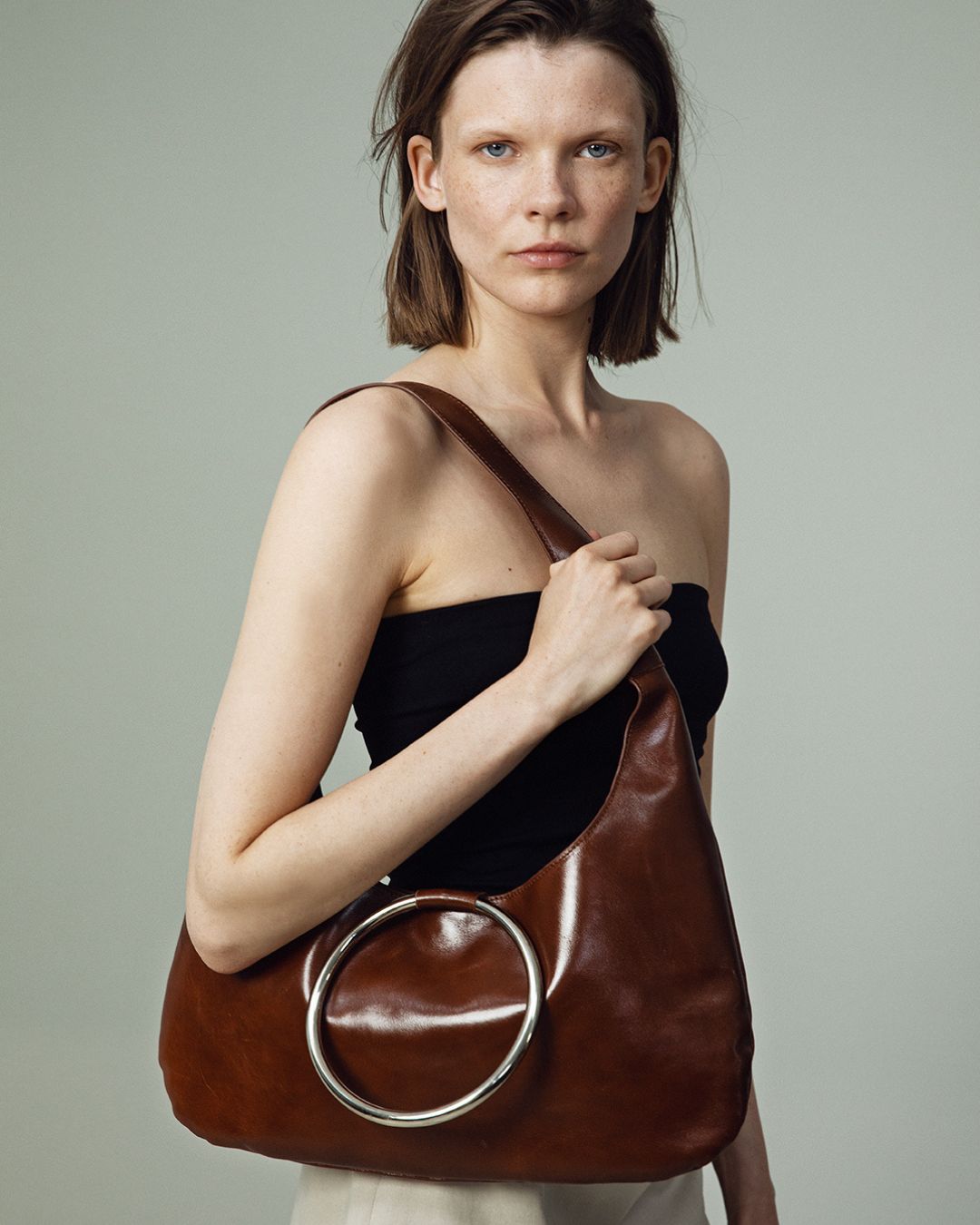 A person holding a KALDA brown shoulder bag with a signature metal circle closure.