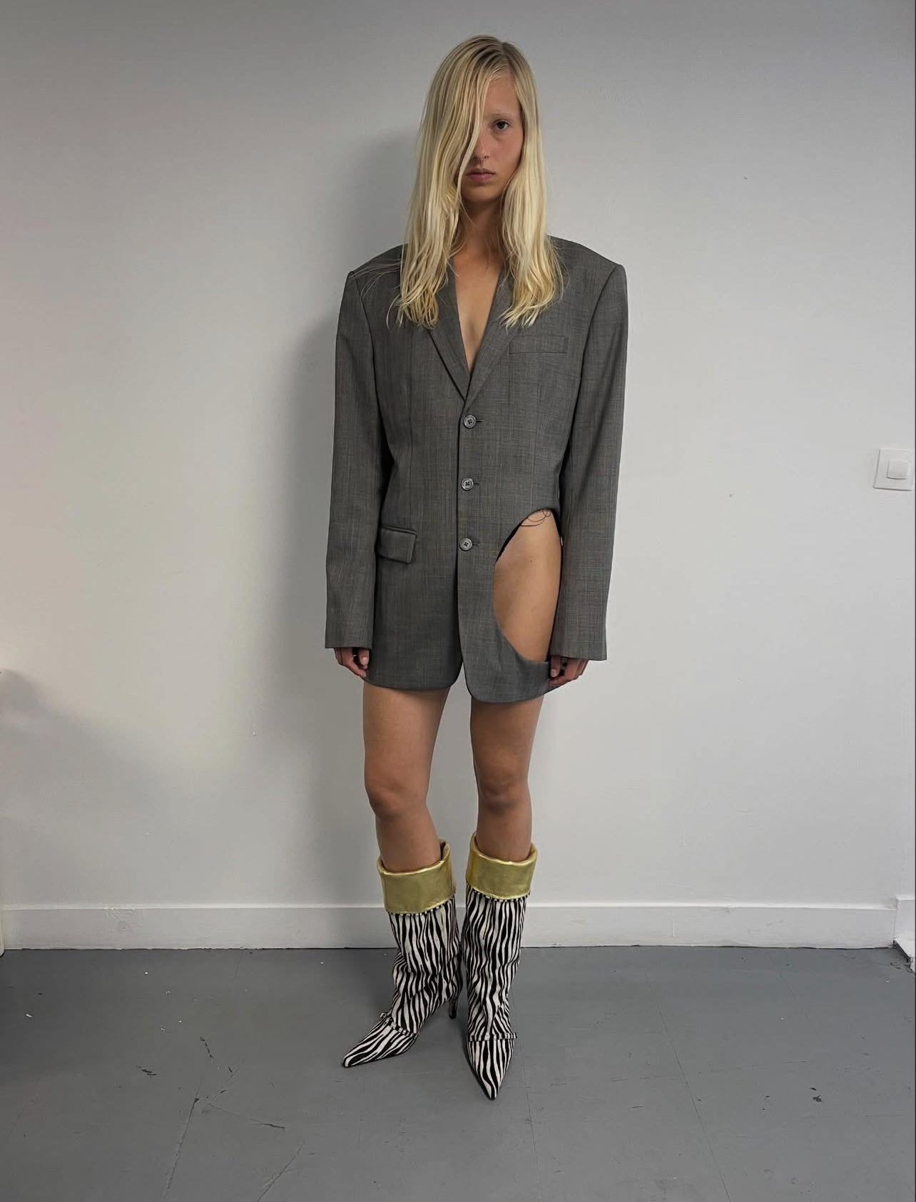 Person wearing a gray blazer over shorts with zebra-striped boots against a plain wall.